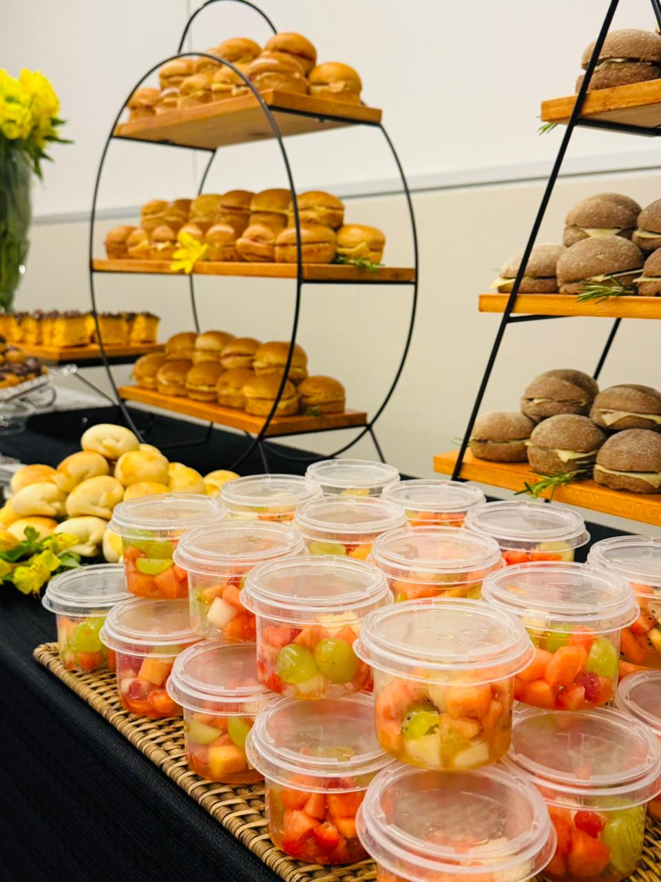 Mesa de coffee break com saladas de frutas individuais e torres de sanduíches